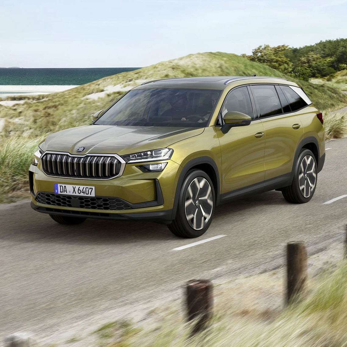 Grüner Škoda Karoq in seitlicher Frontansicht fährt über eine Straße zwischen den Dünen, im Hintergrund das Meer