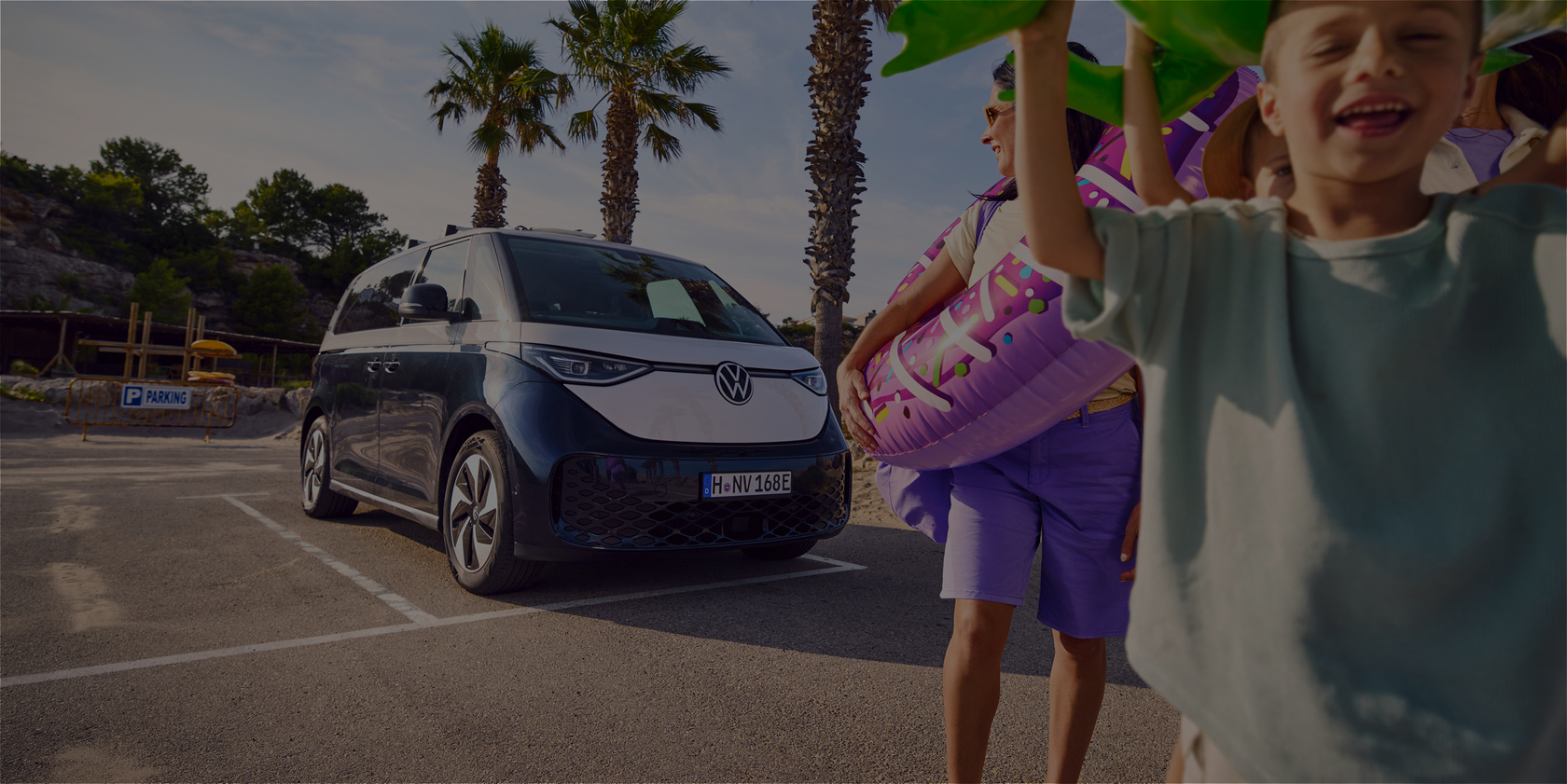 Volkswagen ID. Buzz auf einem Strandparkplatz, im Vordergrund lachende Kinder mit Schwimmreifen, sommerliche Urlaubsstimmung