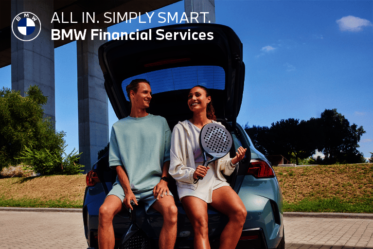 Junges Paar mit Padel-Schlägern lehnt am geöffneten Kofferraum eines BMW – BMW Financial Services Werbung mit dem Slogan 'ALL IN. SIMPLY SMART.