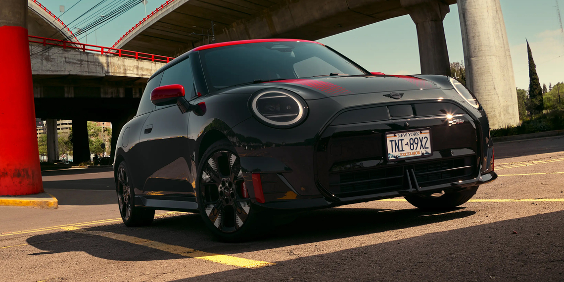 Schwarzer MINI John Cooper Works in Frontansicht mit roten Folierungen und Akzenten, geparkt unter einer Brücke mit rotem Pfeiler sowie roten Leitplanken.