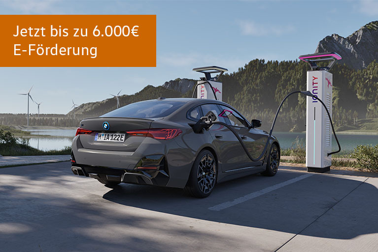 Graues BMW i4 Elektro-Coupé beim Laden an einer Schnellladesäule vor Berg- und Seenlandschaft, mit Hinweis auf bis zu 6.000 Euro E-Förderung.