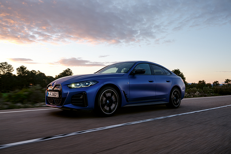 Mattblauer BMW i4 M50 in seitlicher Ansicht. Er fährt auf einer Straße mit ländlichem Panorama, im Hintergrund ein Sonnenuntergang.