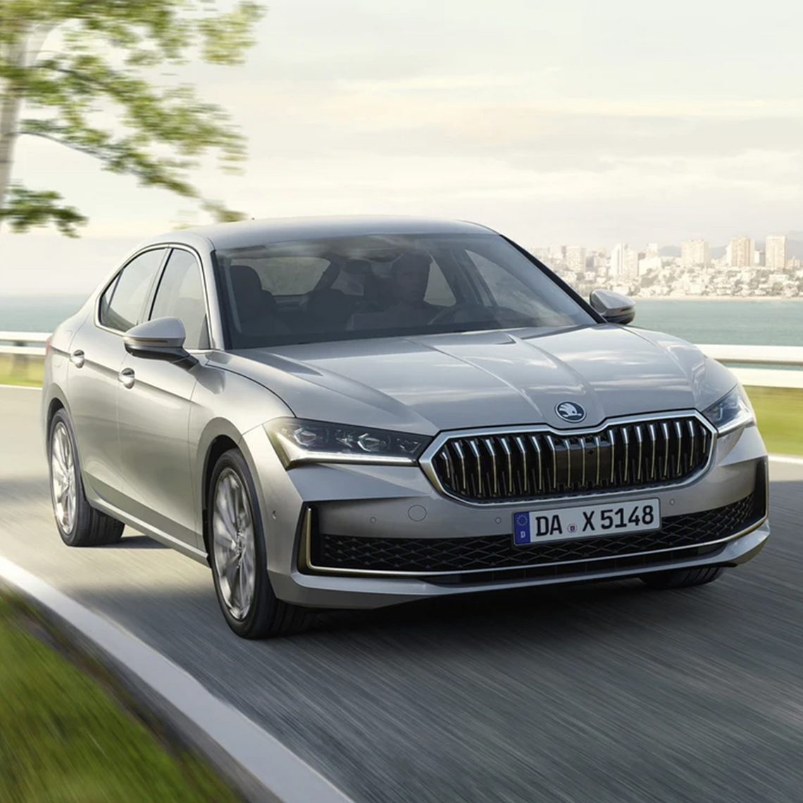 Ein grauer Škoda Superb in Frontansicht fährt auf einer Straße. Im Hintergrund das Meer, dahinter eine weit entfernte Stadt mit Hochhäusern.