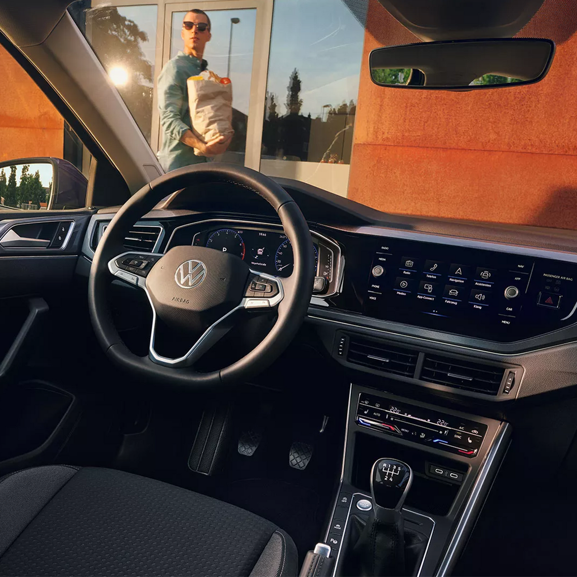 Cockpit eines VW Polo mit Multifunktionslenkrad und großem Infotainmentsystem, durch die Frontscheibe sieht man eine Person mit Einkaufstasche vor Glasfassade.