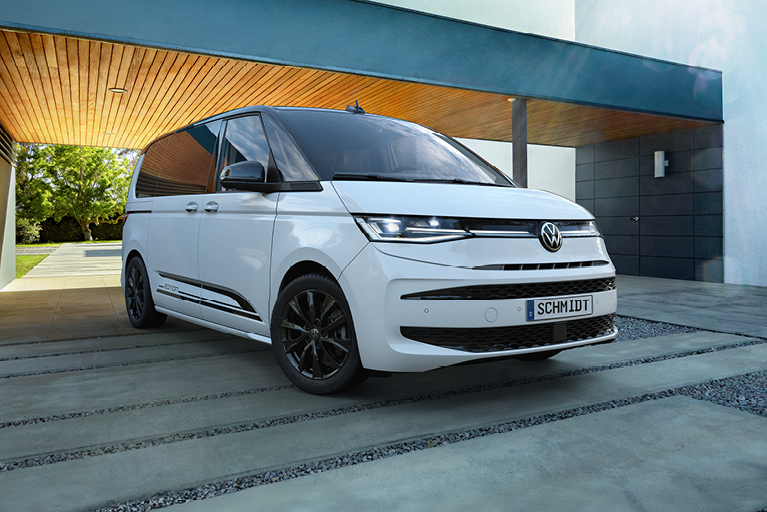 Weißer Volkswagen Multivan in seitlicher Frontansicht mit schwarzen Akzenten und Schriftzug "Edition", geparkt vor der Eingangstür eines modernen Gebäudes.