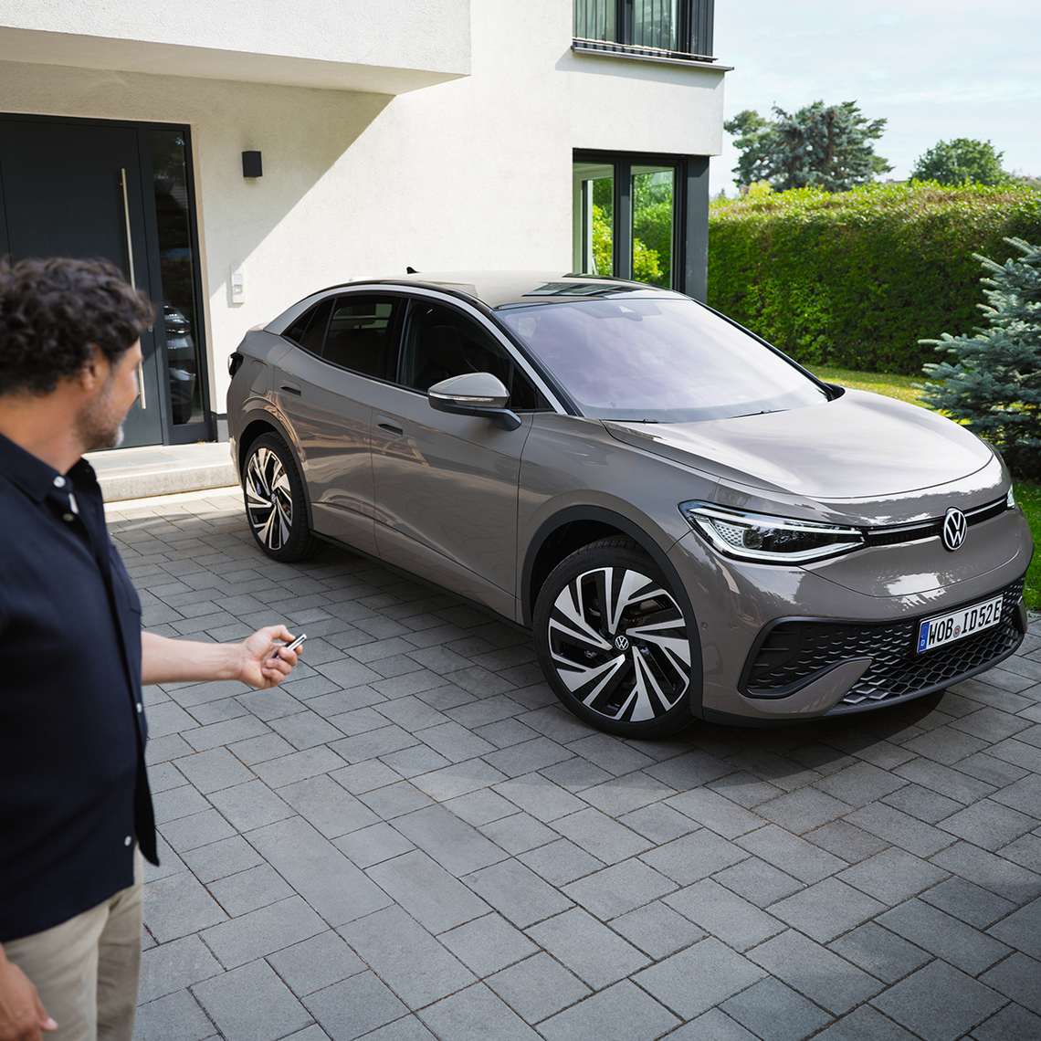 Mann mit Autoschlüssel vor einem geparkten grauen VW ID.5 auf einer Einfahrt vor einem modernen Wohnhaus.