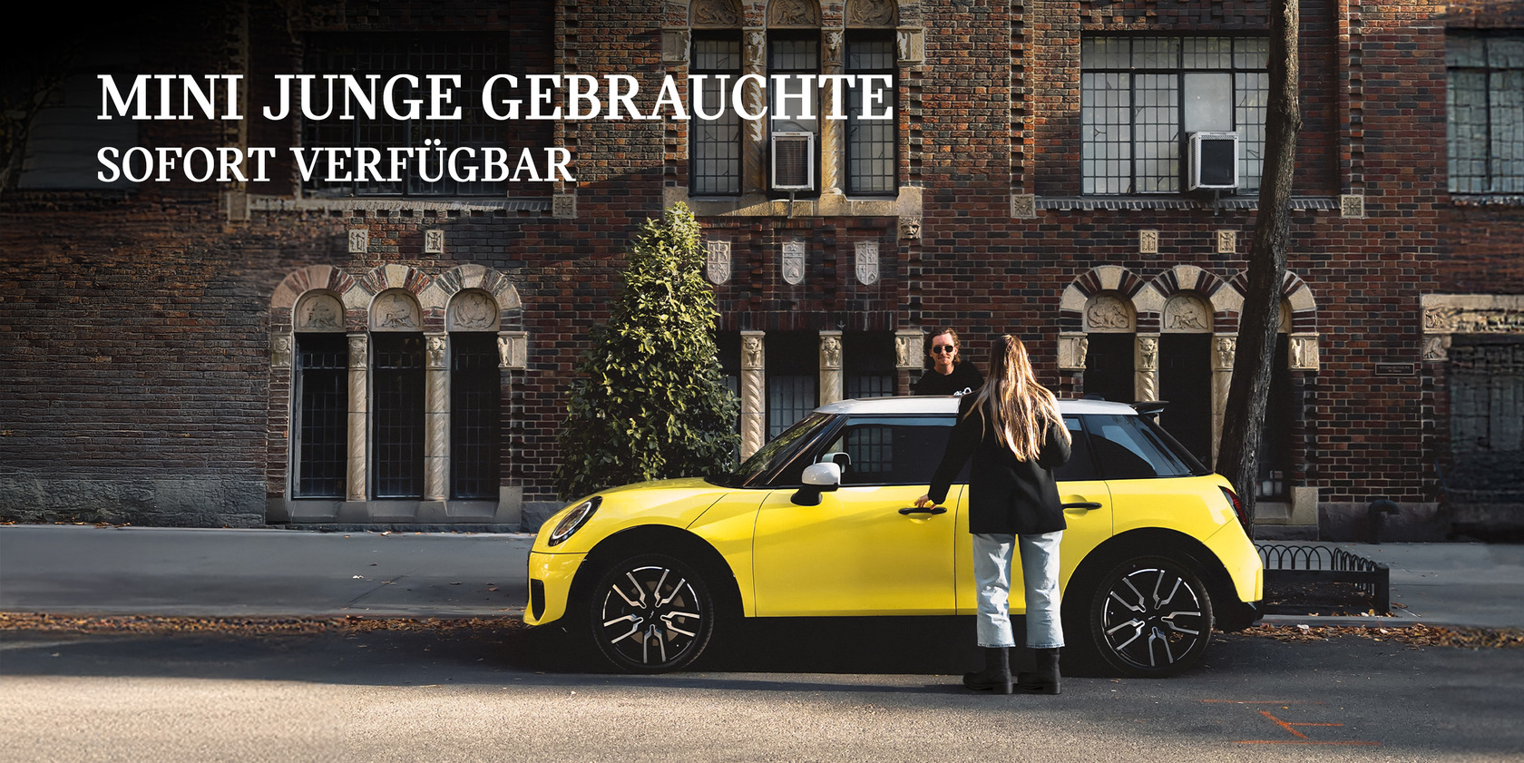 Gelber MINI vor Backsteingebäude in der Stadt mit zwei Personen und dem Schriftzug ‚MINI Junge Gebrauchte sofort verfügbar