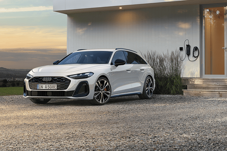 Weißer Audi A5 Avant TFSI e plug-in-hybrid geparkt vor einem modernen Haus bei Sonnenuntergang, mit Ladekabel an der Hauswand – ein stilvoller Kombi in eleganter Umgebung.