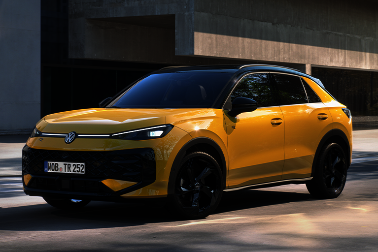 Gelber VW T-Roc R-Line vor einem modernen Betongebäude bei Tageslicht, fotografiert leicht schräg von vorn rechts.