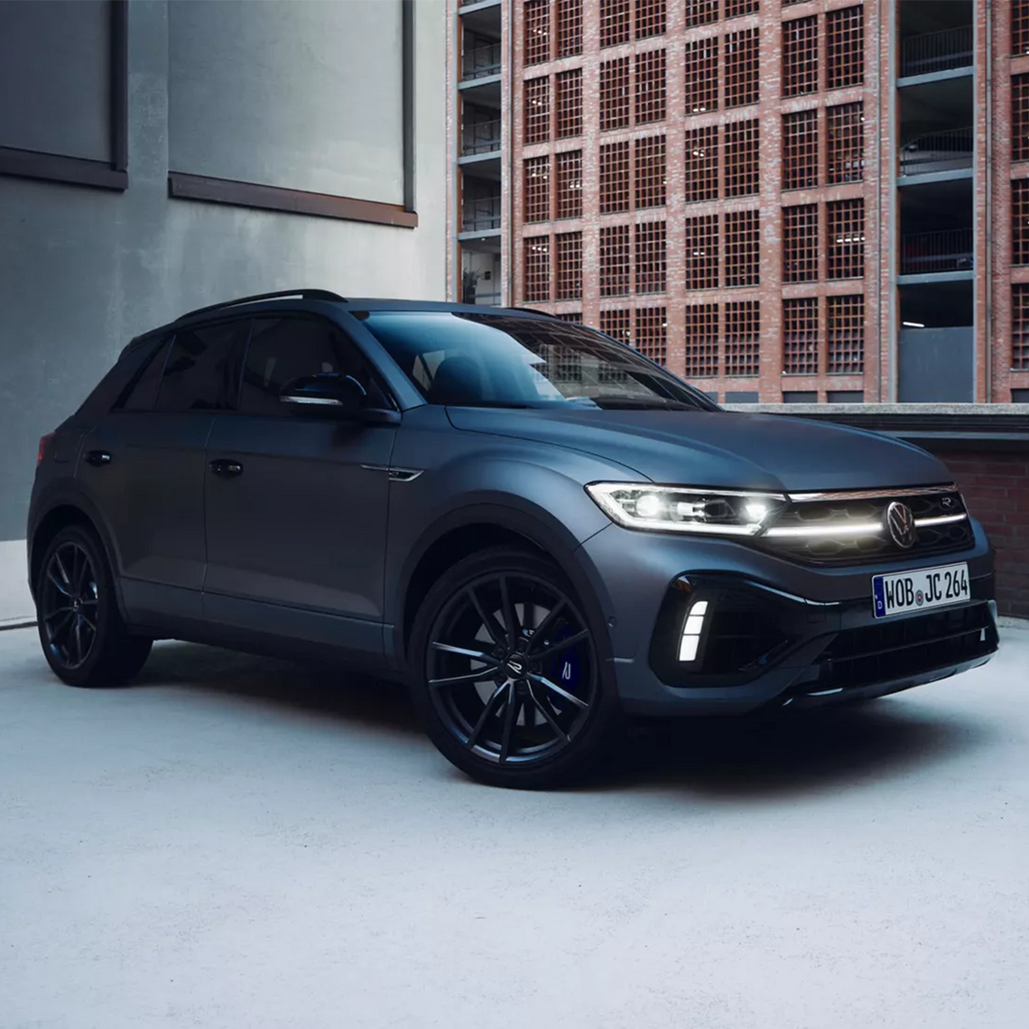 Dunkelgrauer matter VW T-Roc R in Seiten- und Frontansicht gepart vor einem modernen Parkhaus mit rotem Backstein und Gitterfassade.