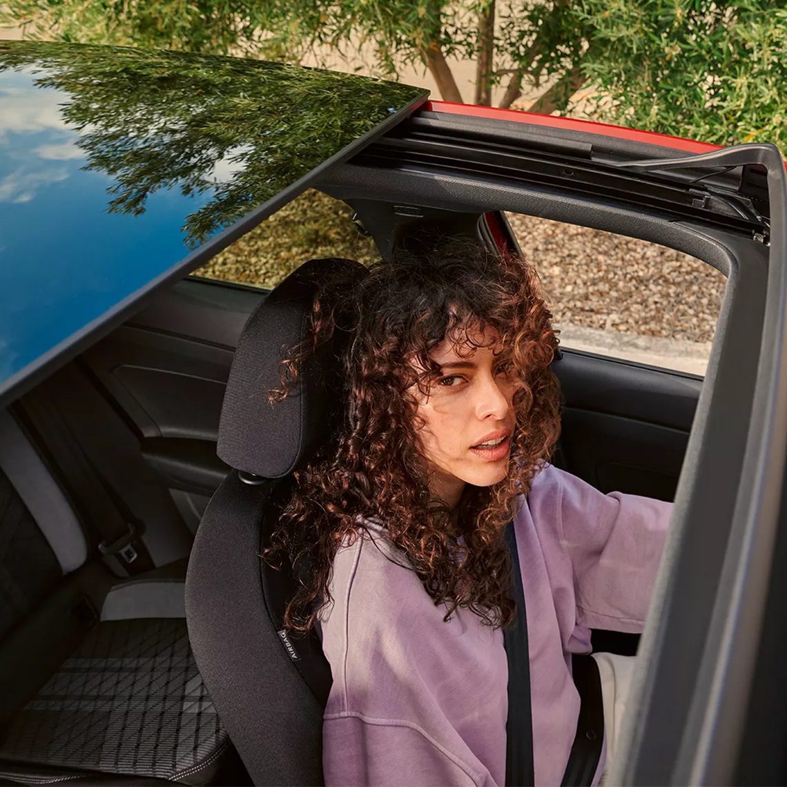 Frau mit lockigen Haaren und lila Pullover sitzt angeschnallt auf dem Fahrersitz eines Autos, fotografiert von oben durch das geöffnete Panoramadach, mit Spiegelung des Himmels und Bäumen auf dem Glas.