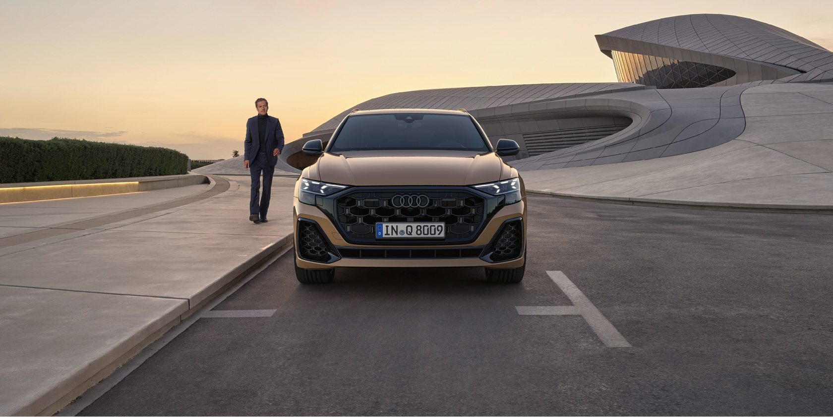 Braun/goldener Audi Q8 bei Sonnenuntergang in Frontalansicht, geparkt vor moderner Architektur. Links neben dem Auto ein Mann gehend auf dem Bürgersteig.