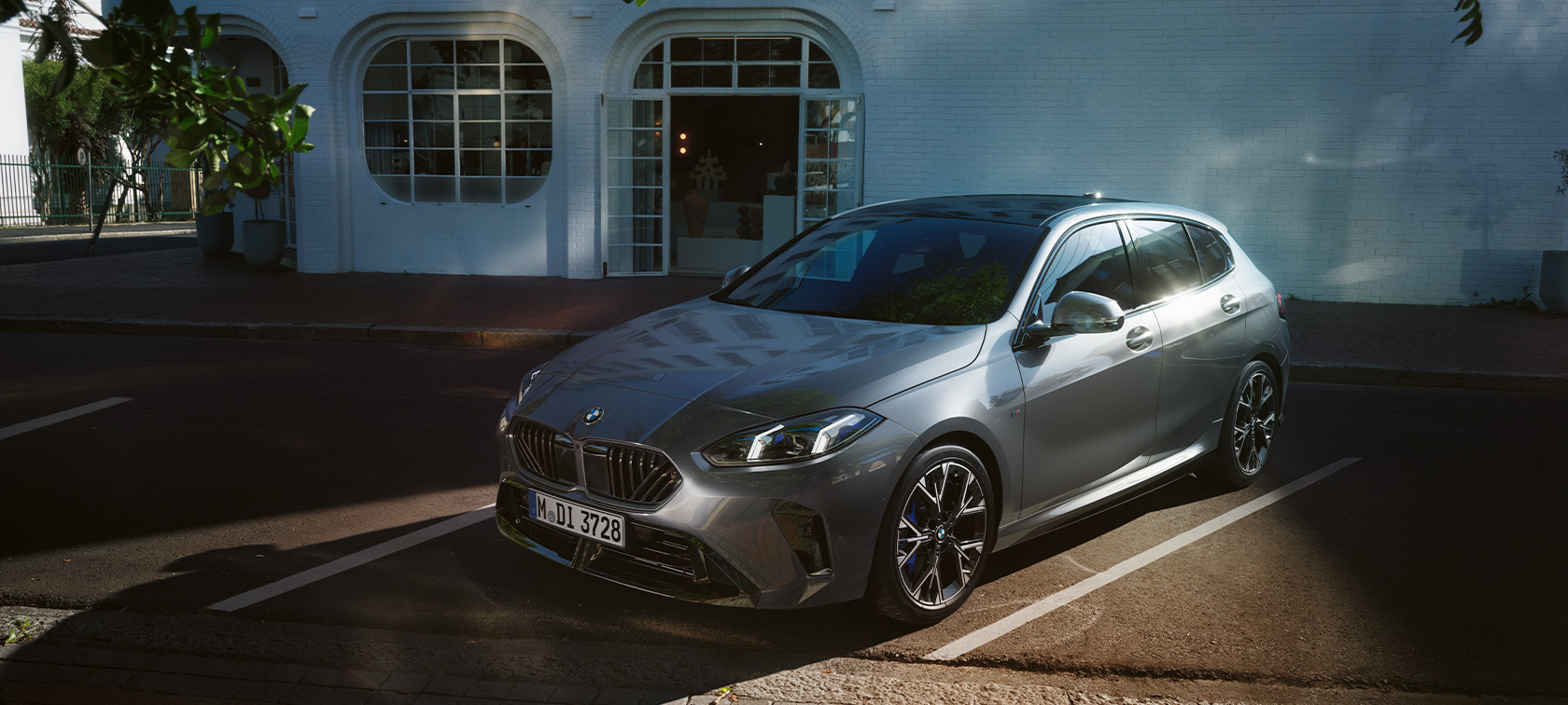 Grauer BMW in Front- und Seitenansicht, geparkt auf einem gekennzeichneten Parkplatz vor einem weißen Gebäude mit geöffneten Flügeltüren.