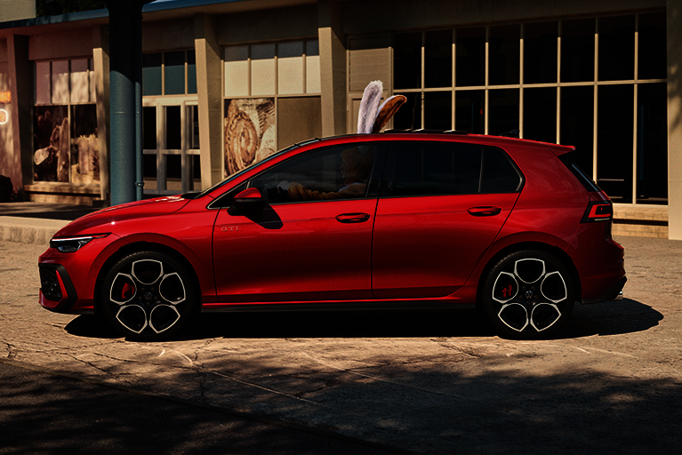 Roter Volkswagen Golf GTI mit sportlichem Design, seitlich fotografiert, mit einem Osterhasen als Fahrerfigur vor urbaner Kulisse.