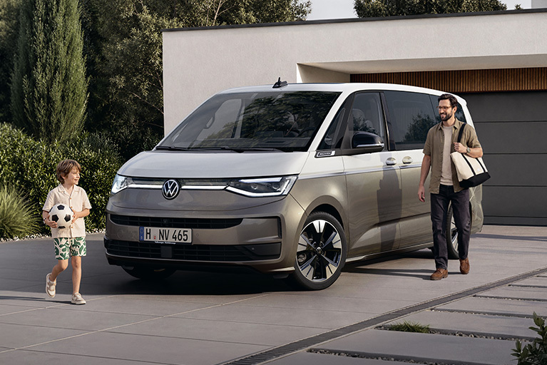 Grauer Volkswagen Multivan vor einem modernen Wohnhaus. Ein Mann mit Tasche und ein Kind mit Fußball stehen neben dem Fahrzeug.