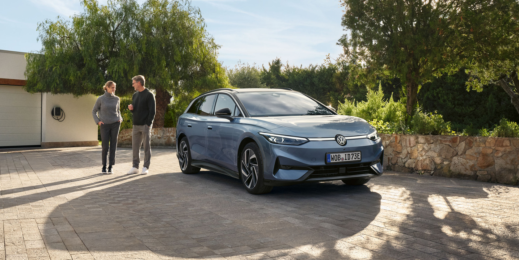 Hellblauer VW ID.7 Tourer in Front- und Seitenansicht geparkt in einer modernen Garage mit Wallbox. Ein Mann und eine Frau stehen neben dem Fahrzeug und schauen sich an. Im Hintergrund blühende Bäume und eine Hecke.