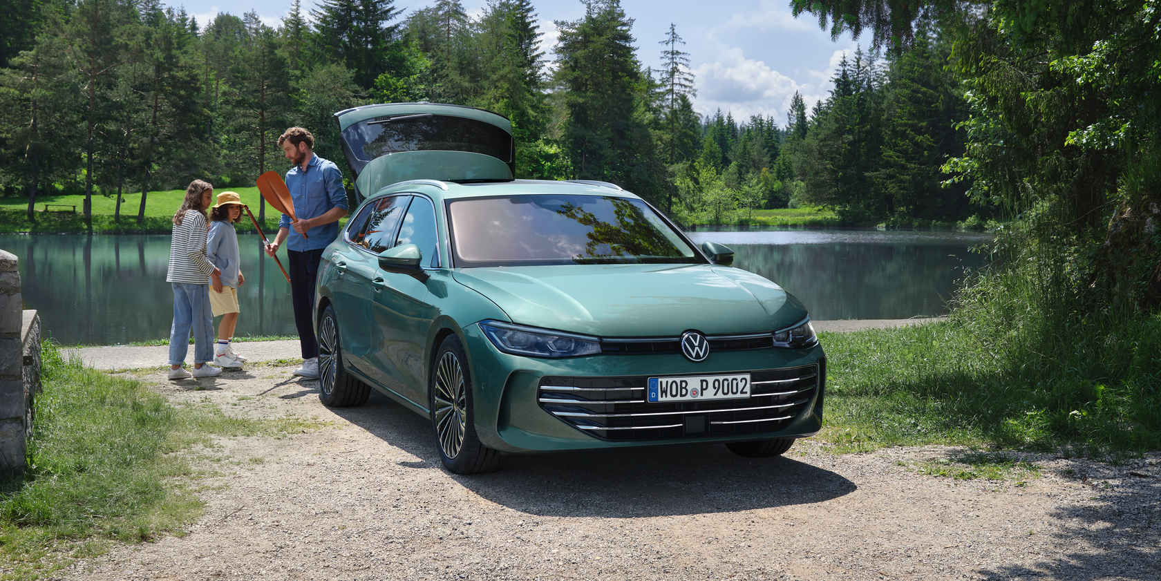 Ein dunkelgrüner VW Passat in Front- und Seitenansicht vor einem blauen See, umgeben von vielen Bäumen und grüner Wiese. Am geöffneten Kofferraum des Fahrzeugs steht ein Mann mit einem Paddel in der Hand und zwei Kinder.