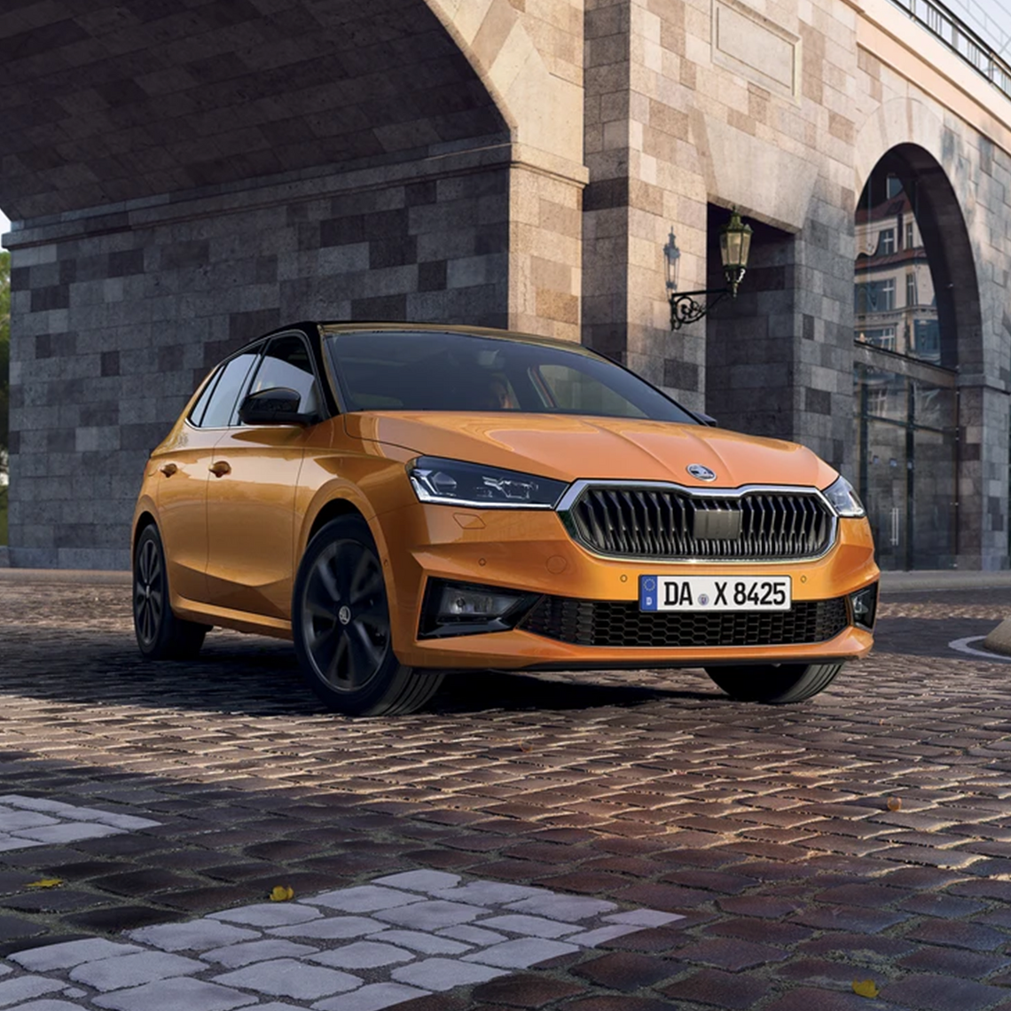 Orangener Škoda Fabia in seitlicher Frontansicht, geparkt in einer städtischen Umgebung auf einem gepflasterten Platz unter einer Brücke