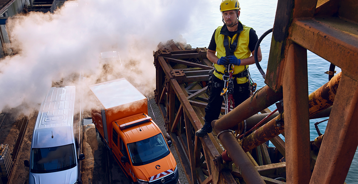 Industriearbeiter in Sicherungsausrüstung steht auf einer Stahlkonstruktion über zwei Volkswagen Nutzfahrzeugen: einem weißen Transporter und einem orangefarbenen Kastenwagen, umgeben von Dampf und Wasser.