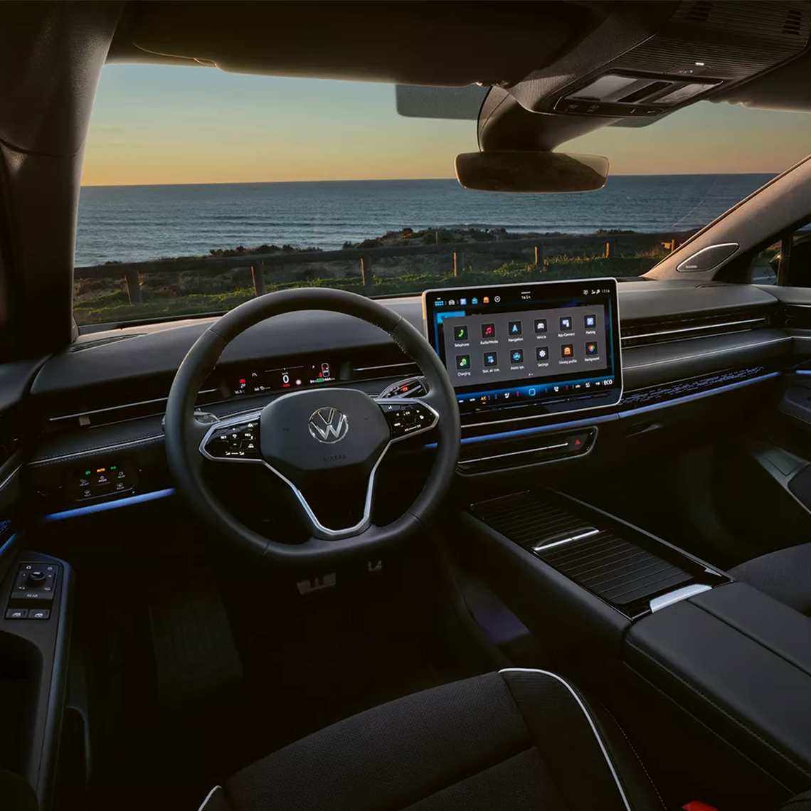 Innenraum des Volkswagen ID.7 mit großem Touchdisplay, digitalem Cockpit und blauer Ambientebeleuchtung. Durch die Frontscheibe sieht man das Meer bei Sonnenuntergang.