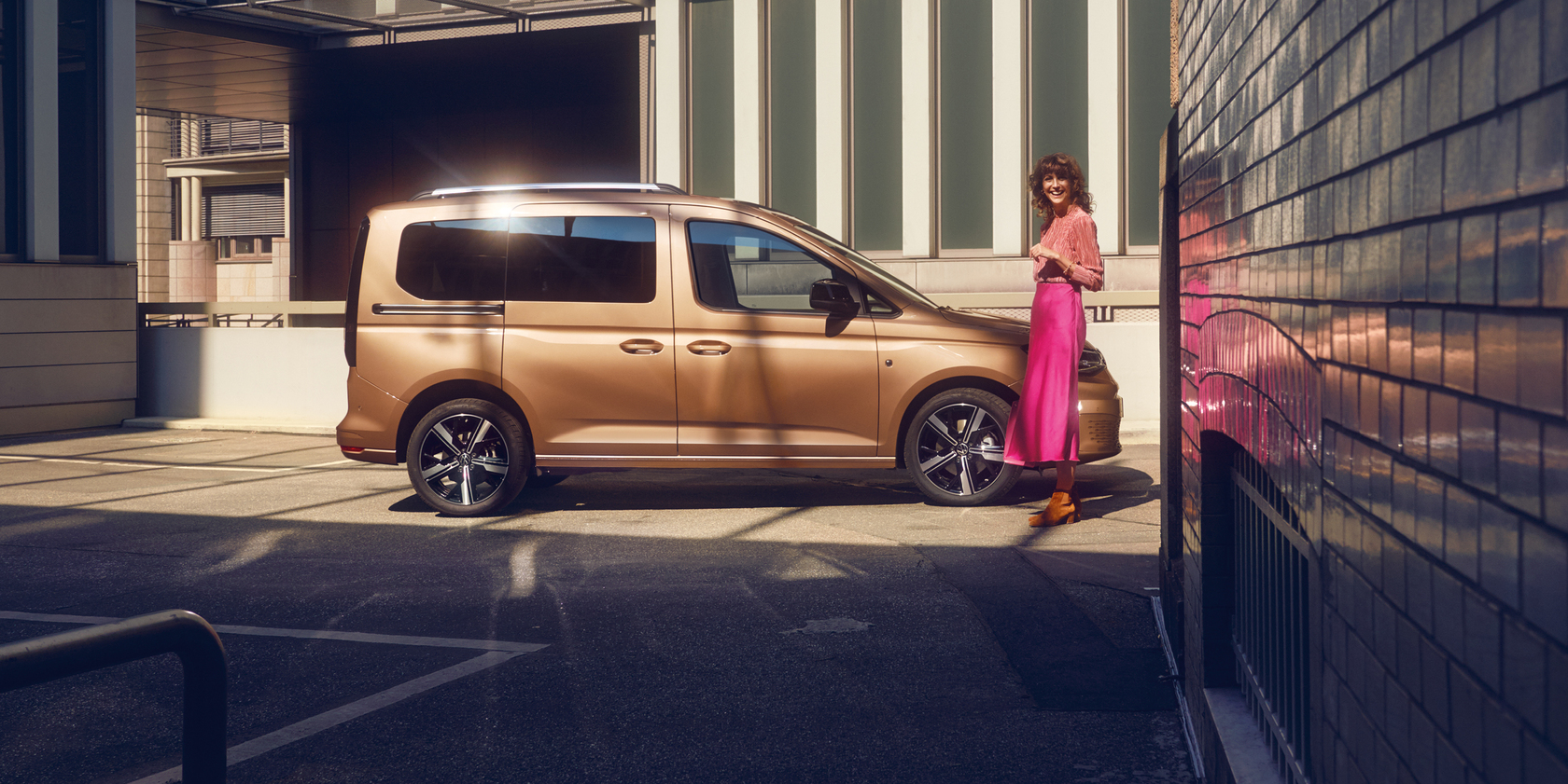 Kupferfarbener Volkswagen Caddy Kombi in Seitenansicht geparkt auf einem Parkdeck, umgeben von Gebäuden. Am vorderen Teil des Autos steht eine Frau, die in die Kamera lächelt.