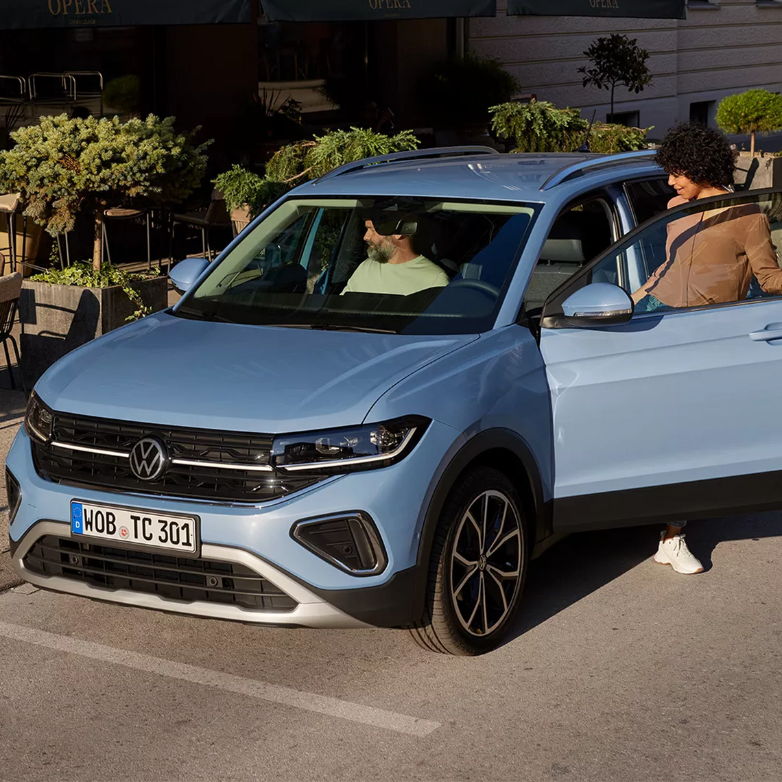 Hellblauer Volkswagen T-Cross in Frontansicht geparkt vor einem Café auf einem sonnigen Parkplatz, eine Frau steigt auf der Fahrerseite ein, ein Mann sitzt bereits auf dem Beifahrersitz.