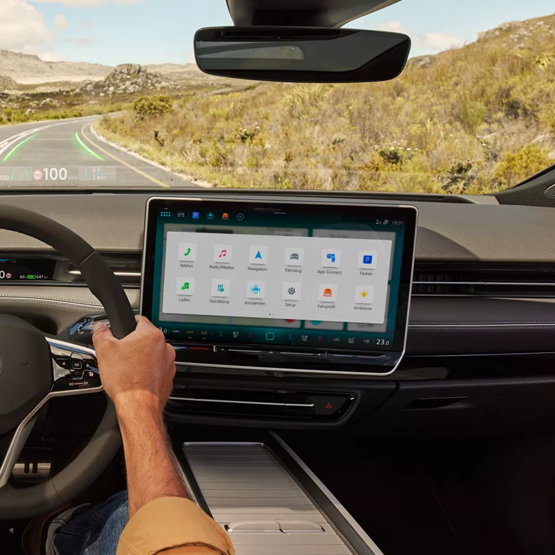 Cockpitansicht eines VW Modells mit großem Touchdisplay und Head-Up-Display. Eine Fahrerhand am Lenkrad während der Fahrt durch eine felsige Landschaft