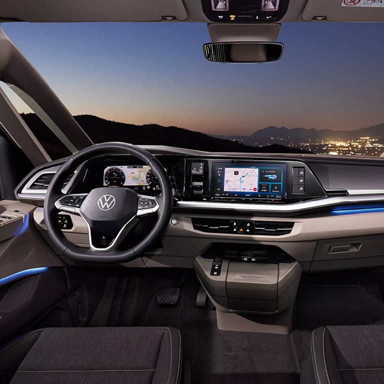 Cockpit eines VW California Beach mit Multifunktionslenkrad, Infotainment-Touchdisplay, Automatikgetriebe und blauer Ambientebeleuchtung. Durch die Frontscheibe sieht man eine beleuchtete Stadt zwischen den Bergen bei Nacht.
