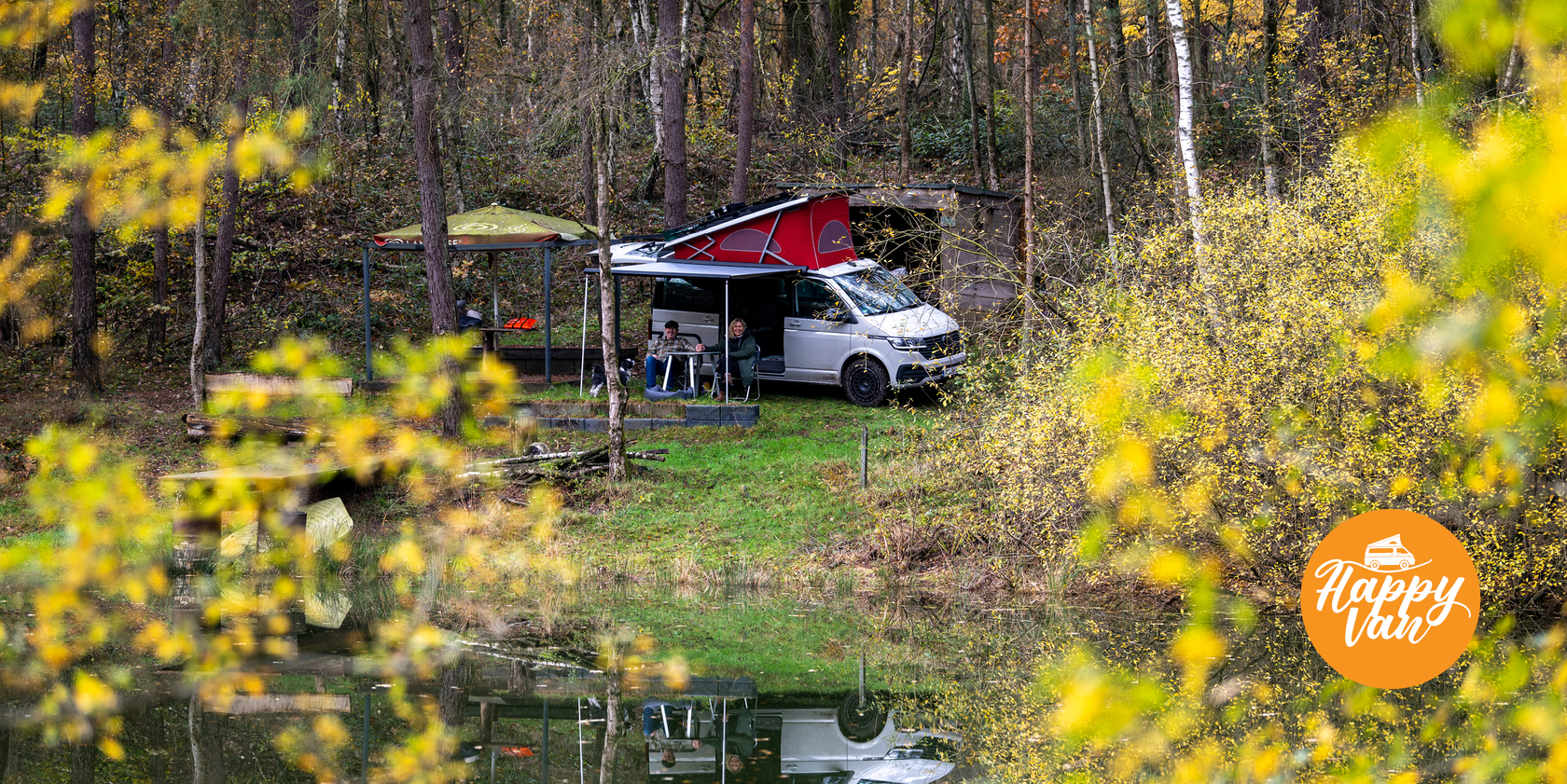VW Californa mit aufgeklapptem Aufstelldach steht am Waldrand neben einem kleinen See. Zwei Personen sitzen unter dem ausgefahrenen Sonnendach, umgeben von buntem Herbstlaub. Unten rechts im Bild das Happy Van Logo