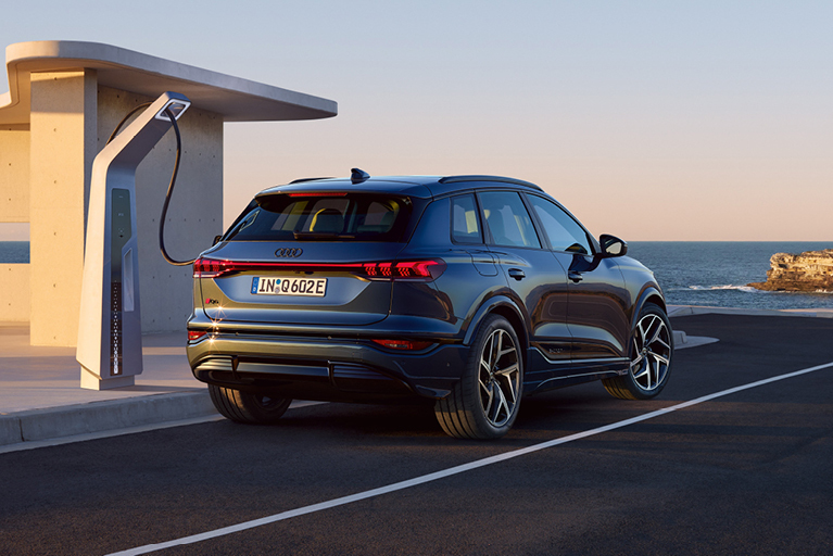 Ein Audi Q4 e-tron in dunkelgrau angeschlossen an einer Ladesäule. Im Hintergrund die Küste mit Blick auf das Meer. 