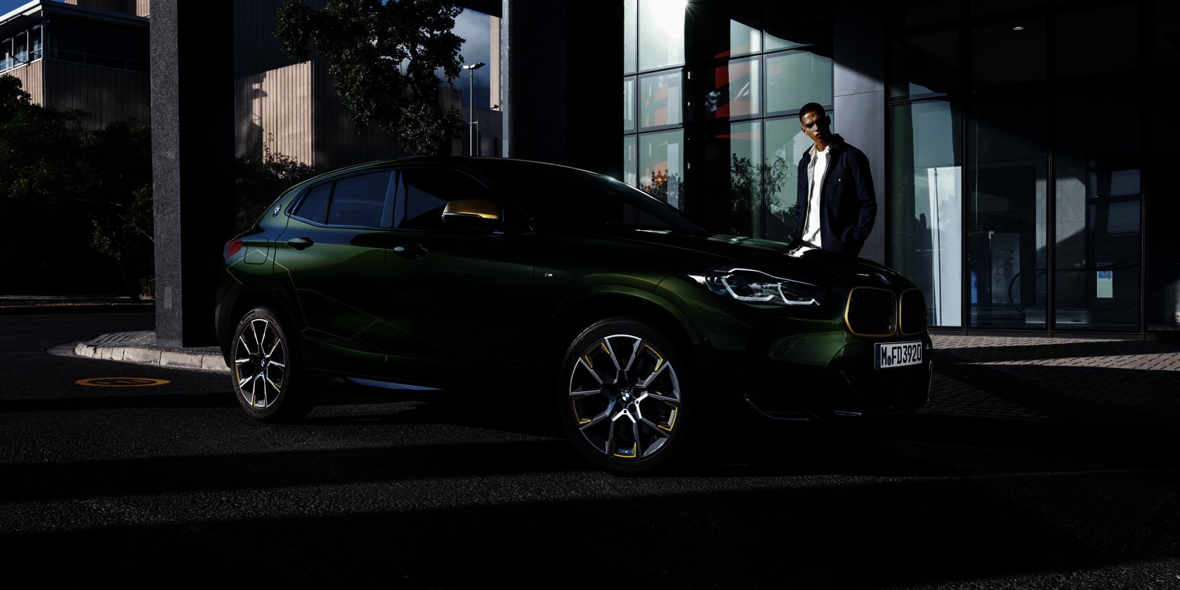 BMW X2 in auffälligem grün-goldenem Design, geparkt vor einem modernen Bürogebäude, mit stilvoll gekleideter Person im Hintergrund – sportlich-urbanes Lifestyle-Statement.