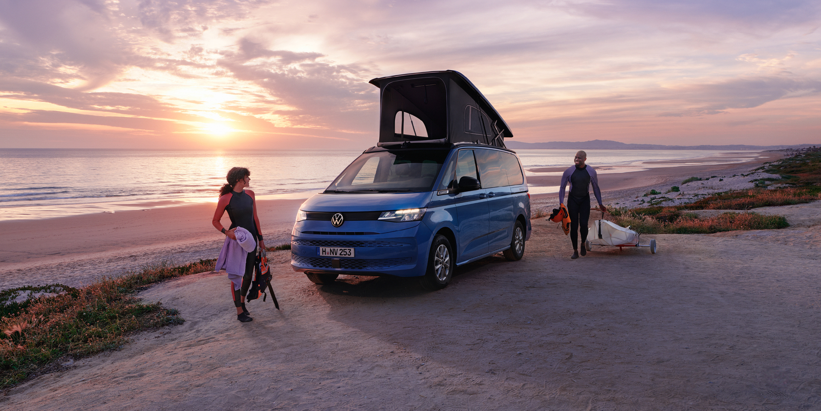 Blauer VW California Campervan mit ausgeklapptem Aufstelldach steht am Strand bei Sonnenuntergang. Zwei Personen in Neoprenanzügen laufen in Richtung Fahrzeug.