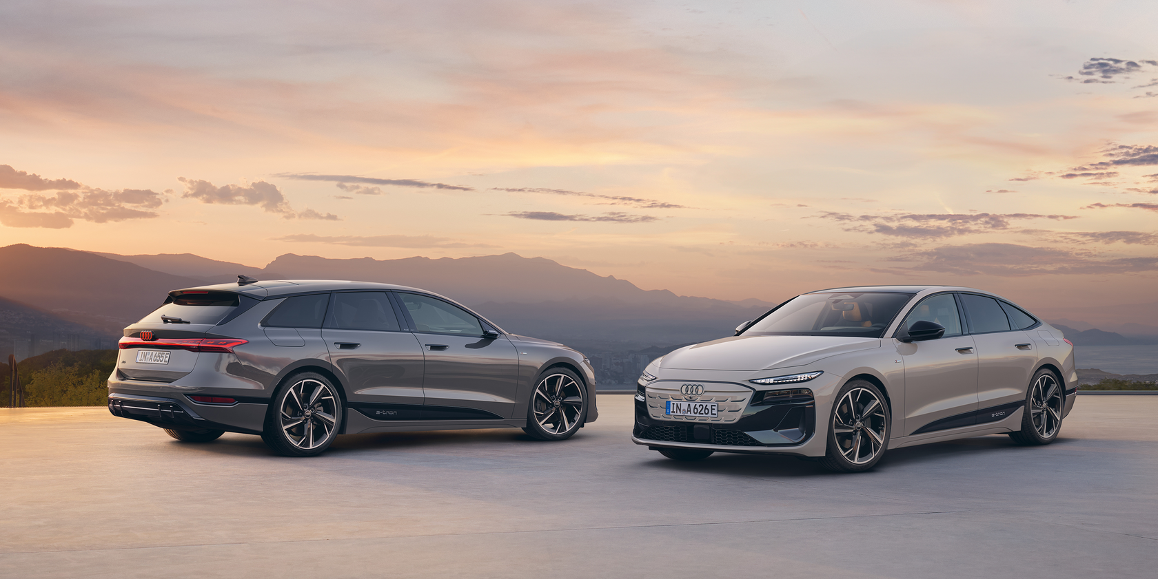 Zwei Audi A6 e-tron Modelle stehen in entgegengesetzter Richtung zueinander: Links ein dunkelgrauer Kombi in Seiten- und Heckansicht. Links eine beige Limousine in Front- und Seitenansicht. Im Hintergrund Berge und ein Sonnenuntergang.