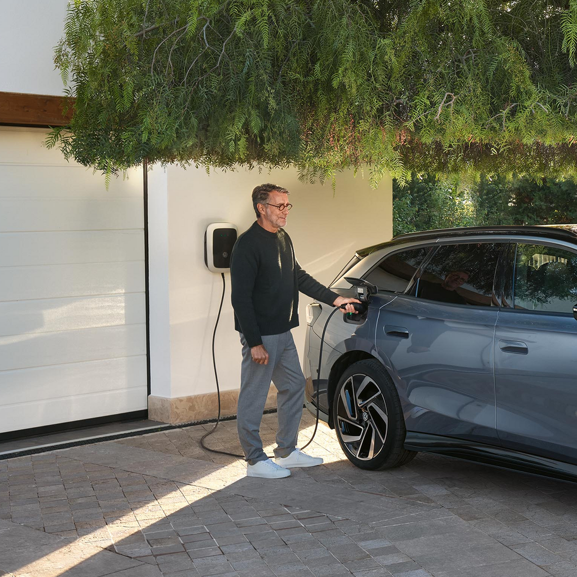 Mann lädt einen hellblauen VW ID.7 an einer Wallbox vor einer Garage an einem modernen Haus