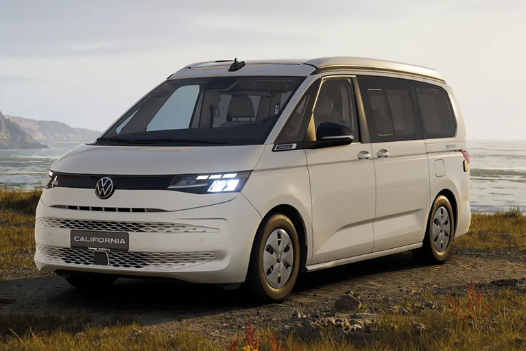 Weißer Volkswagen California Beach Camper am Wasser geparkt, mit Blick auf eine felsige Küste bei Sonnenuntergang.