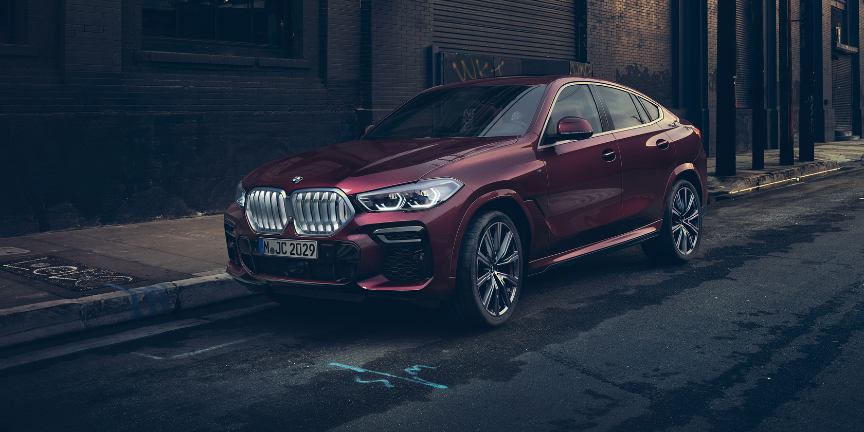Dunkelroter BMW X6 in seitlicher Frontansicht, geparkt am Straßenrand vor einem dunklen Gebäude.